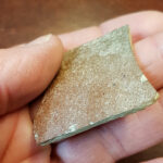 Das Bild zeigt die Scherbe. Sie wird schräg in der Hand gehalten. Dadurch sieht man leichte Rillen auf der Oberfläche der Keramik.